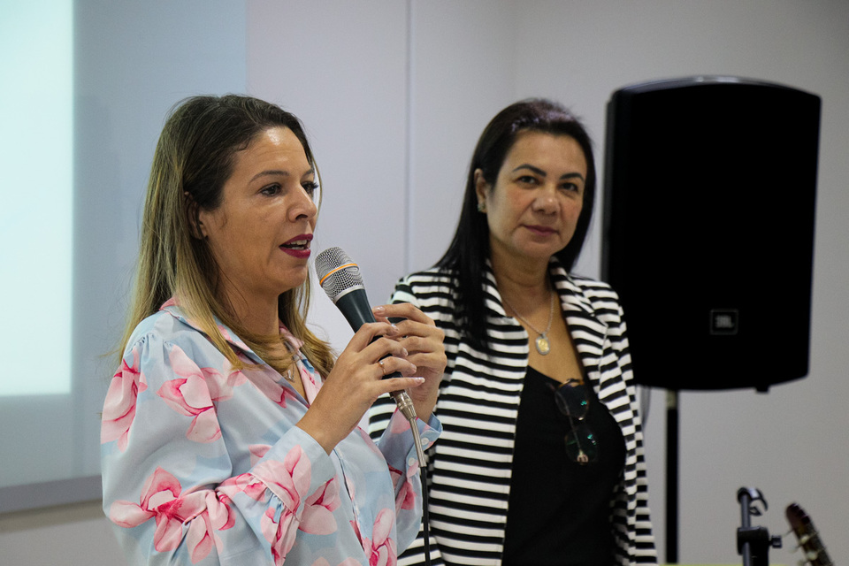 mulher loira com microfone na mão ao lado de uma mulher de cabelo preto. Amas estão em pé